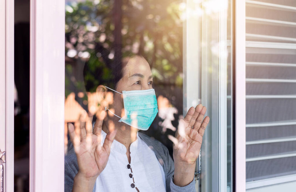 Пожилые азиатские женщины в маске с руками, касающимися окна, сидят дома во время коронавируса и эпидемии ковида-19 или пандемии, концепция социального дистанцирования - Фото, изображение