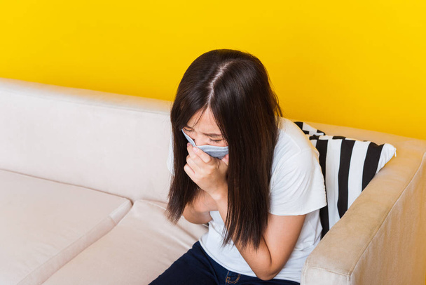 Asian young woman sitting on a sofa wearing surgical protection face mask hygiene against coronavirus and her sneeze hand close mouth, studio shot isolated on yellow background with copy space - Photo, image