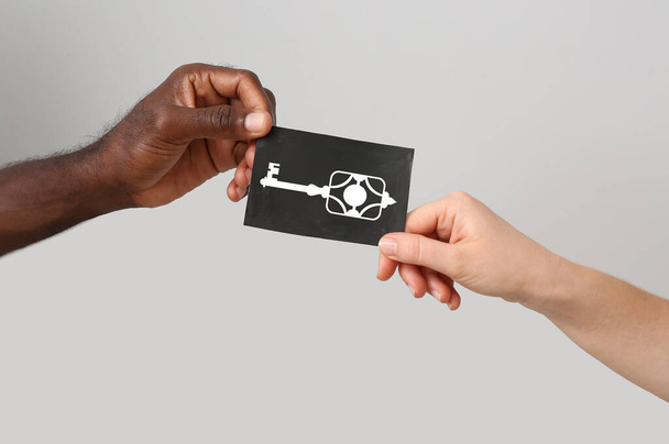 Hände einer kaukasischen Frau und eines afroamerikanischen Mannes mit gezogenem Schlüssel auf Papierbogen vor grauem Hintergrund - Foto, Bild
