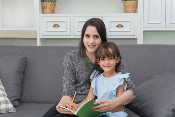Young mother teaching daughter holding book sitting on sofa at home, family leisure activities. - Foto, Bild