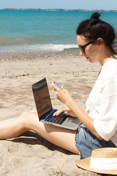 Jeune femme utilisant un ordinateur portable sur une plage. Communication sur le mode de vie des vacances. Il tient une carte bancaire entre ses mains. Le concept d'achat en ligne ou d'achat en ligne - Photo, image