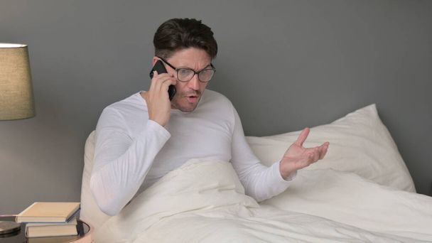 Dühös középkorú férfi beszél okostelefonon az ágyban - Fotó, kép