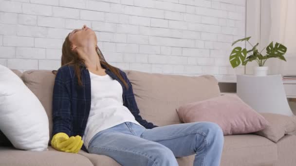 Jeune femme fatiguée dans des gants de protection en caoutchouc assis sur le canapé, se détendre après le travail de nettoyage de la maison à la maison - Séquence, vidéo