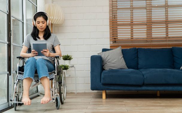 Mulher asiática deficiente feliz sentada em uma cadeira de rodas e trabalhando com tablet em casa, o conceito de tecnologias para a conveniência de pessoas com deficiência. - Foto, Imagem