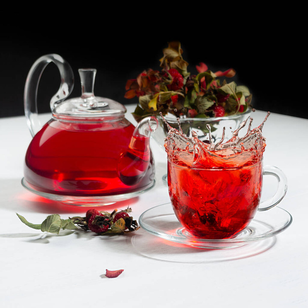 Autumn evening teatime with splash in red herbal tea cup, transparent tea pot with hawthorn berries, rose hip, dry leaves on white wood table, black wall, square. - Photo, Image