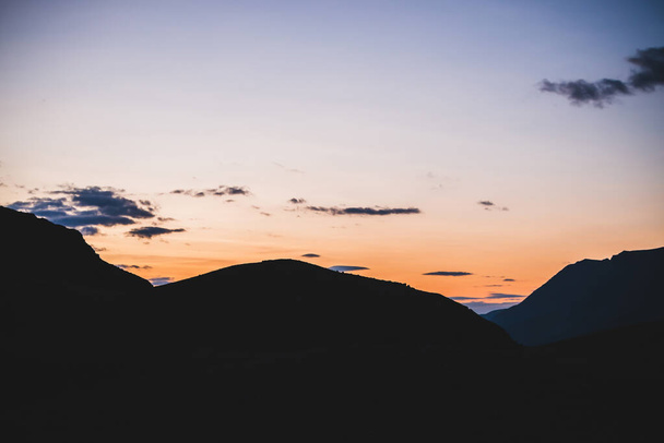 Atmospheric minimalist alpine landscape to big mountain silhouettes in sunset. Giant darker mountains under dawn sky with clouds. Vivid orange sundown. Wonderful highland scenery to multicolor dawn. - Foto, Imagen