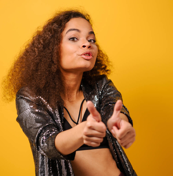 Thumbs up happy Afro American girl with beautiful hairstyle posing smile looking side away with hand on hips wearing shiny black jacket and black top on yellow background - Zdjęcie, obraz