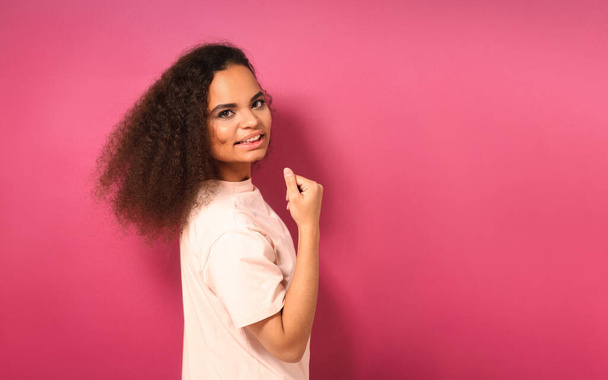 Beautiful young African American girl half turned looking positively at camera wearing peachy t-shirt showing her muscles isolated on pink background. Beauty concept - Fotografie, Obrázek