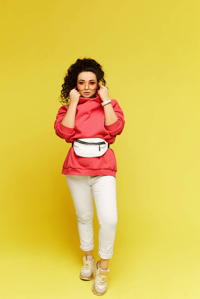 Young woman in a pink hoodie, white pants and sunglasses posing over yellow background. Model in sportswear with beltbag on the yellow background - Fotoğraf, Görsel