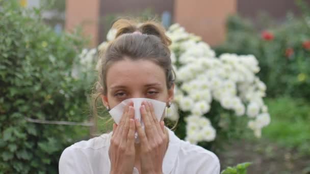 Girl sneezes into a napkin, because she is allergic to flowering during spring time on white chrysanthemums on background, hypersensitivity concept - Footage, Video
