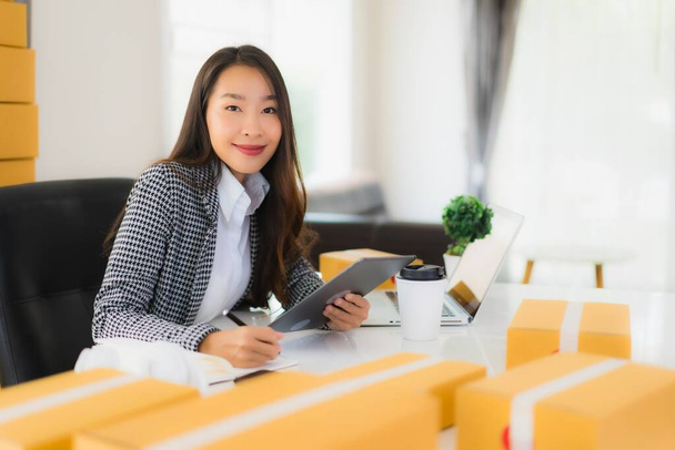 Portrait beautiful young asian business woman work from home with laptop mobile phone with cardboard box ready for shipping online shopping - Photo, Image