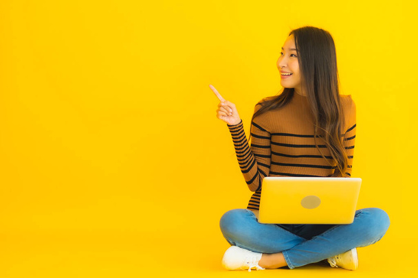 肖像美しい若いアジアの女性は黄色の隔離された背景にラップトップやコンピュータを使用するために床に座っている - 写真・画像