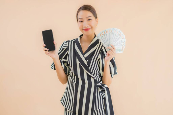 Portrait beautiful young asian woman with cash and smart mobile phone on brown background - Φωτογραφία, εικόνα