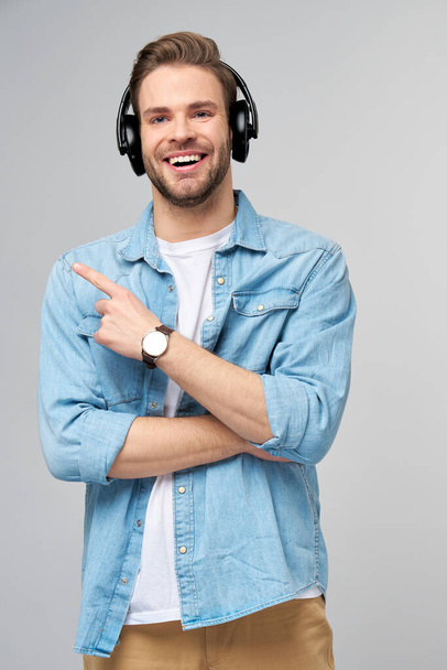 Happy young handsome man in jeans shirt pointing away standing against grey background wearing big headphones - Fotó, kép