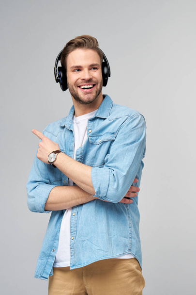 Feliz jovem bonito homem de camisa jeans apontando de pé contra fundo cinza usando grandes fones de ouvido - Foto, Imagem