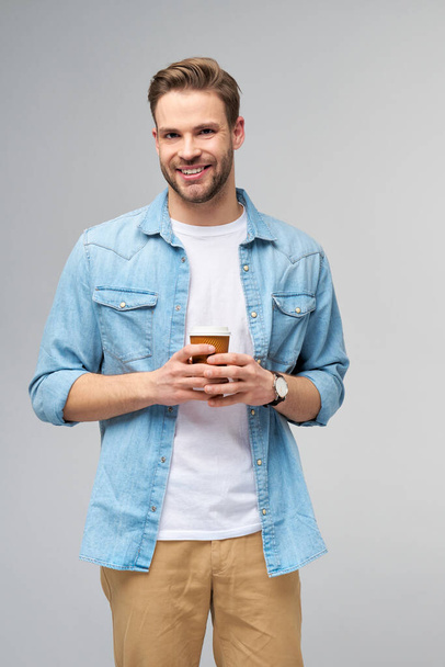 Portrait of young handsome caucasian man in jeans shirt over light background holding cup of coffee to go - Zdjęcie, obraz
