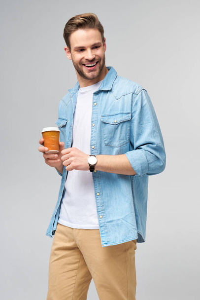 Retrato de joven guapo hombre caucásico en jeans camisa sobre fondo claro sosteniendo taza de café para llevar - Foto, imagen