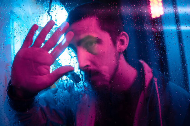 Beautiful cinematic portrait of stylish man in neon lighted window with rain drops. Bright neoned colors. Caucasian model, musician outdoors. Youth culture, scary mist style and music concept. - Фото, зображення