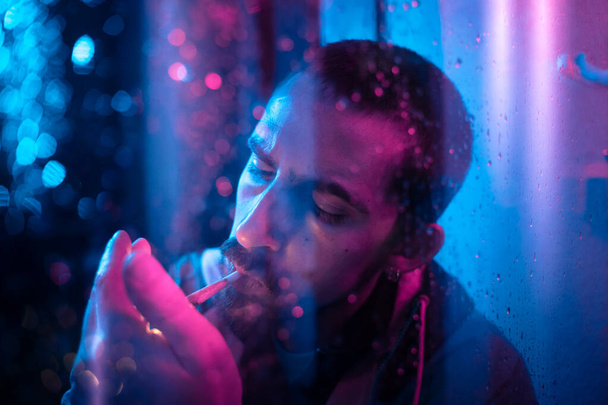 Beautiful cinematic portrait of stylish man in neon lighted window with rain drops. Bright neoned colors. Caucasian model, musician outdoors. Youth culture, scary mist style and music concept. - 写真・画像