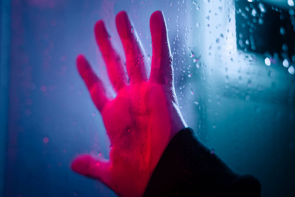 Dramatic hand in neon lighted window with rain drops. Bright neoned colors. Scary and horror concept. - Photo, Image