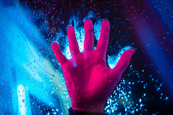 Dramatic hand in neon lighted window with rain drops. Bright neoned colors. Scary and horror concept. - Photo, image