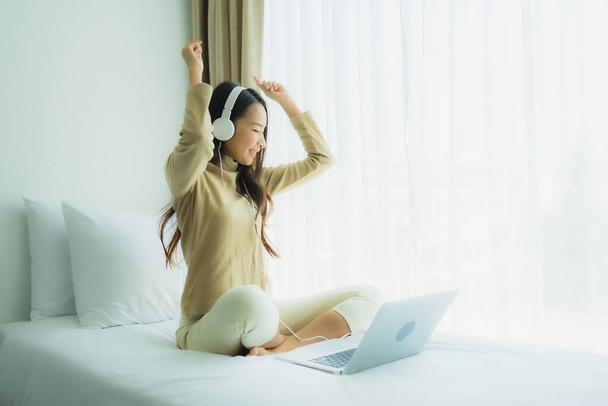 Young asian woman using laptop with headphone for listen music in bedroom interior - 写真・画像