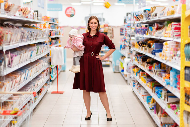 Ostoksilla kaupassa. Nuori nainen hymyilee ja pitää vauvaa sylissään, kun hän poseeraa supermarketin käytävällä. Sisätiloissa. Käsite ostoksia ja kulutustottumukset. - Valokuva, kuva