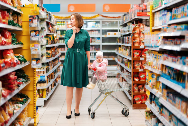Marketten alışveriş. Genç düşünceli bir anne içinde bebek olan bir market arabasını yuvarlıyor. Süpermarket raflarına bak. Alışveriş ve tüketim kavramı. - Fotoğraf, Görsel
