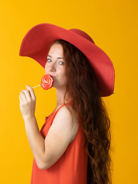 beautiful woman with long hair in a big red hat and lollipop on a yellow background - Fotoğraf, Görsel