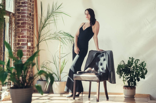 Hermosa modelo de chica vestida con ropa negra de pie cerca del sillón en el interior de estilo loft. - Foto, Imagen