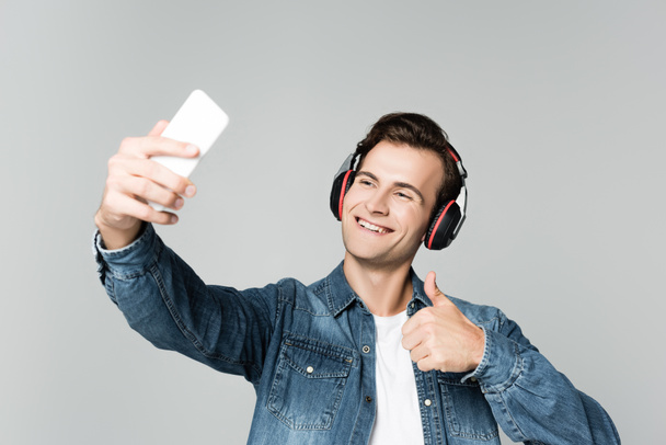 Veselý muž ve sluchátkách ukazuje jako při užívání selfie s chytrým telefonem izolované na šedé - Fotografie, Obrázek