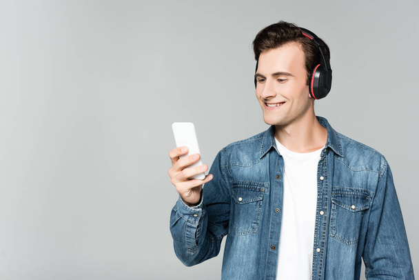 Cheerful man in denim jacket using smartphone and headphones isolated on grey - Photo, Image