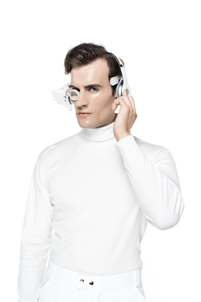 Cyborg in weißer Kleidung, digitaler Augenlinse und Kopfhörer blickt isoliert auf weiße Kamera - Foto, Bild