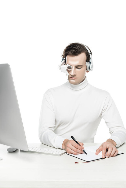 Cyborg-Mann mit Kopfhörer und digitaler Augenlinse schreibt auf Notizbuch in der Nähe von Computer auf Tisch isoliert auf weiß - Foto, Bild