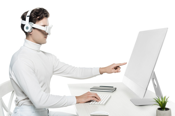 Kyborg ve sluchátkách a oční čočce ukazuje na počítač v blízkosti notebooku a umístit na rozmazané popředí izolované na bílém - Fotografie, Obrázek