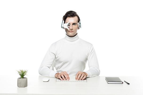 Kyborg ve sluchátkách a oční čočce pomocí počítačové klávesnice poblíž rostlin a notebooku izolovaných na bílém - Fotografie, Obrázek