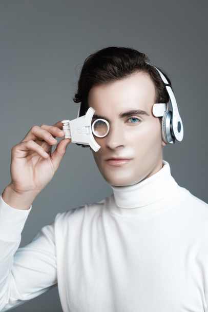 Brunette cyborg in headphones holding digital eye lens isolated on grey - Photo, Image