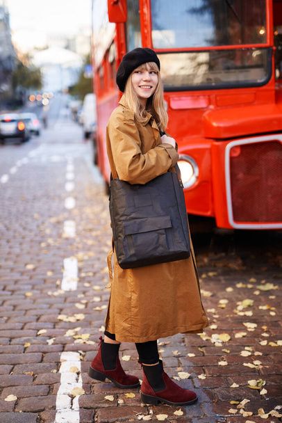 Lächelndes blondes Mädchen in schwarzer Baskenmütze und orangefarbenem Trench auf der Stadtstraße mit rotem Bus. Positives Denken oder Londoner Abenteuerkonzept - Foto, Bild