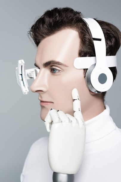 Cyborg hombre con auriculares y dedo de mano artificial cerca de la mejilla aislado en gris - Foto, Imagen