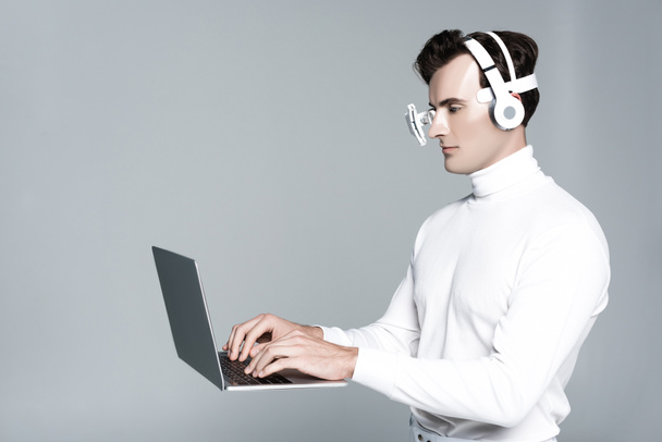 Cyborg in headphones and eye lens using laptop in air isolated on grey - Photo, Image