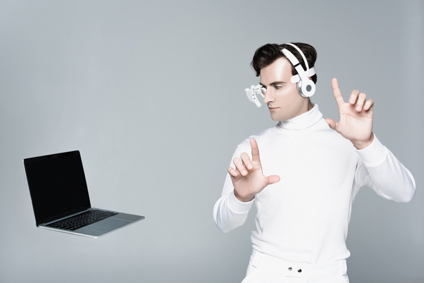 Cyborg in hoofdtelefoon raakt iets aan en kijkt naar laptop met leeg scherm in lucht geïsoleerd op grijs - Foto, afbeelding