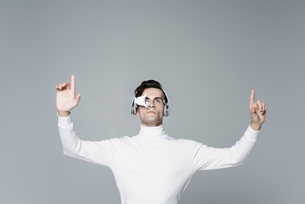 Brunette cyborg man in digital eye lens and headphones looking away while using something isolated on grey - Photo, Image