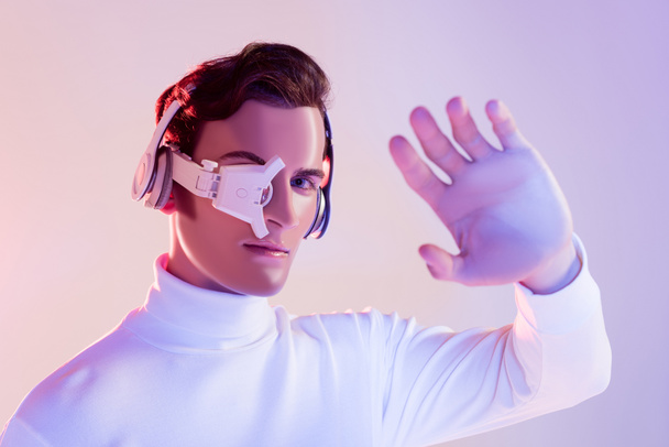 Cyborg in digital eye lens and headphones looking at camera near hand on blurred foreground on purple background - Photo, Image