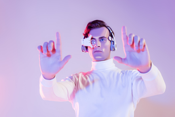 Morena cyborg em lente de olho digital e fones de ouvido usando algo em primeiro plano borrado no fundo roxo - Foto, Imagem