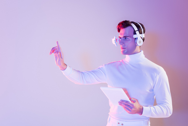 Cyborg in headphones holding digital tablet and using something on purple background - Photo, Image