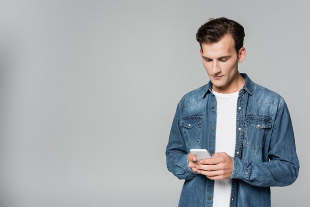 Young man in denim jacket using smartphone isolated on grey - 写真・画像