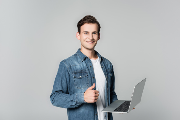 Smiling man showing thumb up while holding laptop isolated on grey - Фото, изображение