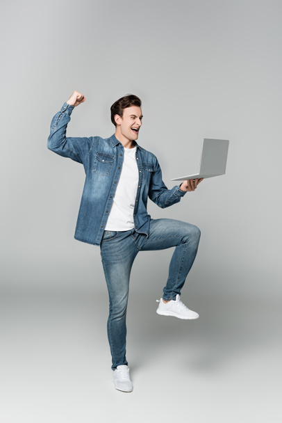 Homme joyeux montrant geste ouais tout en tenant ordinateur portable sur fond gris - Photo, image