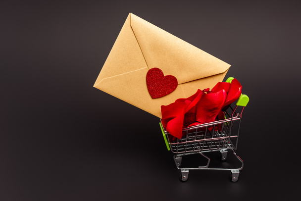 shopping cart with rose petals and envelope isolated on black - Photo, Image