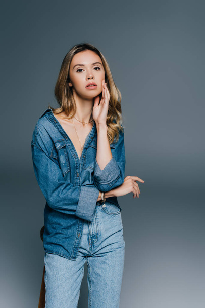 sensual young woman in denim shirt and jeans touching face while looking at camera on grey - Photo, image